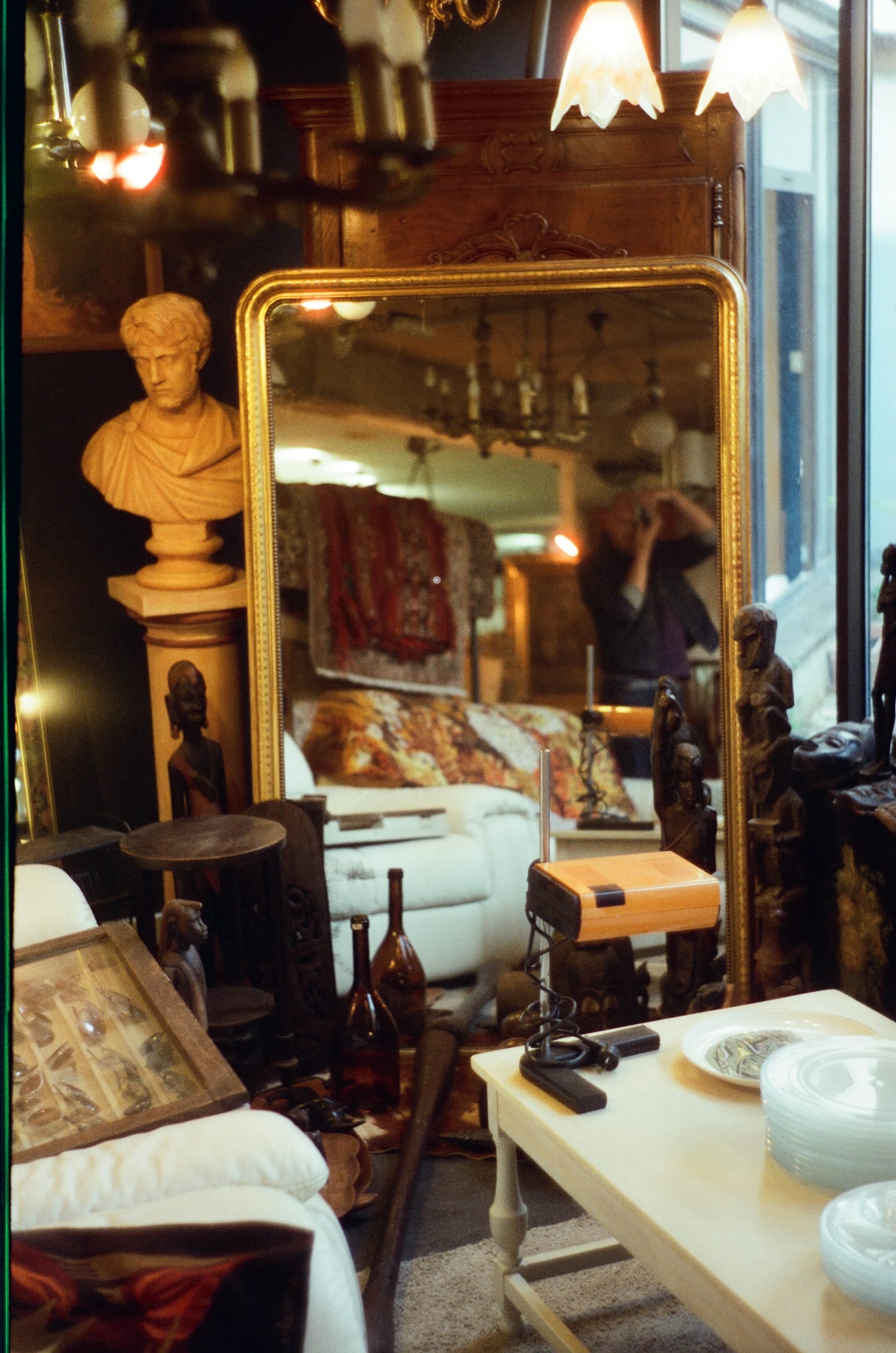 Intérieur de brocante avec miroir doré reflétant le photographe, buste romain et lustres, argentique couleur