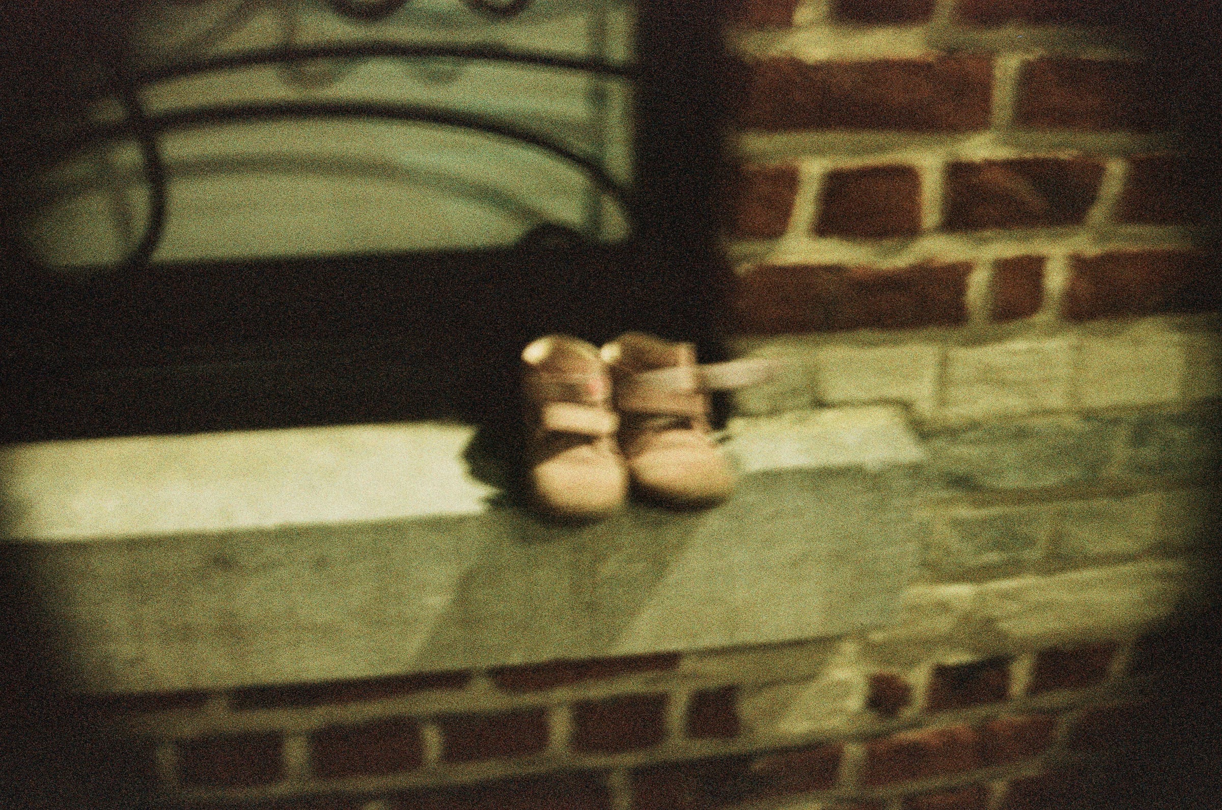 Petites chaussures d'enfant en cuir posées sur un rebord en pierre devant un mur de briques rouges, photographie argentique d'objet trouvé