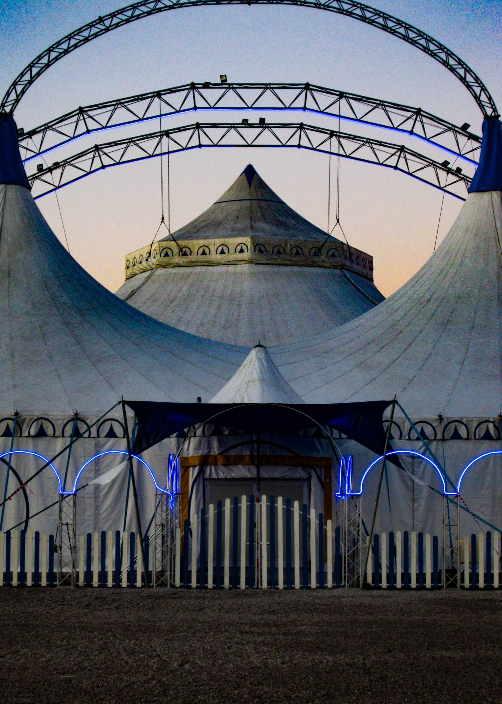 Chapiteau de cirque encadré par des arcs courbes de néon bleu au crépuscule, photographie argentique structurelle et symétrique