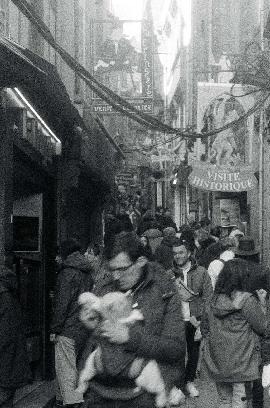 Rue médiévale bondée en France avec silhouettes floues en mouvement et enseignes historiques, argentique noir et blanc