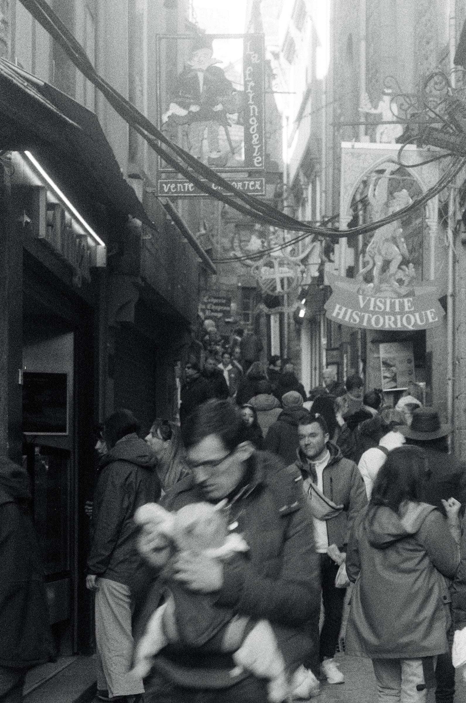 Rue médiévale bondée en France avec silhouettes floues en mouvement et enseignes historiques, argentique noir et blanc
