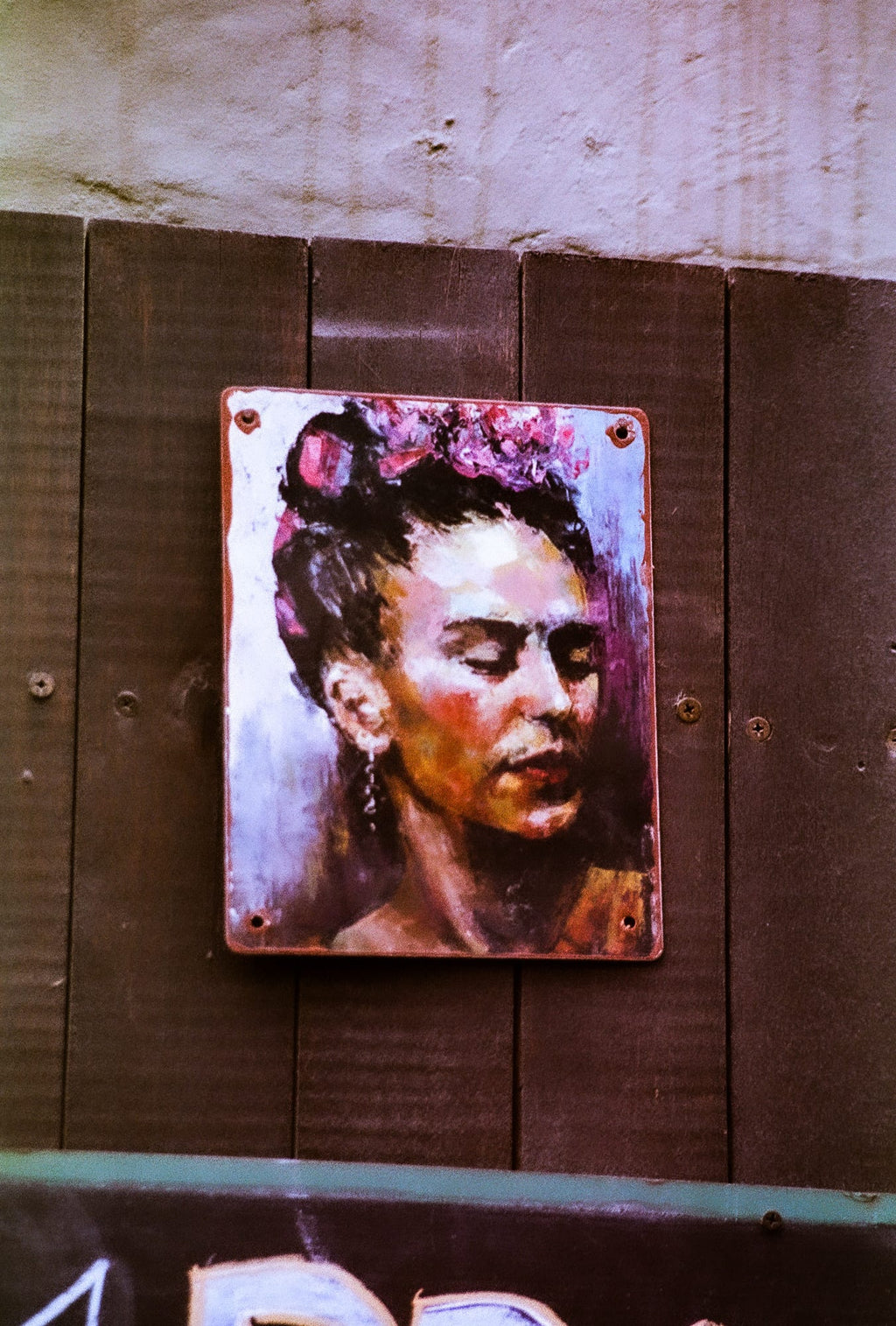 Portrait peint de Frida Kahlo sur une petite plaque métallique fixée sur des planches en bois sombre, détail street art argentique couleur
