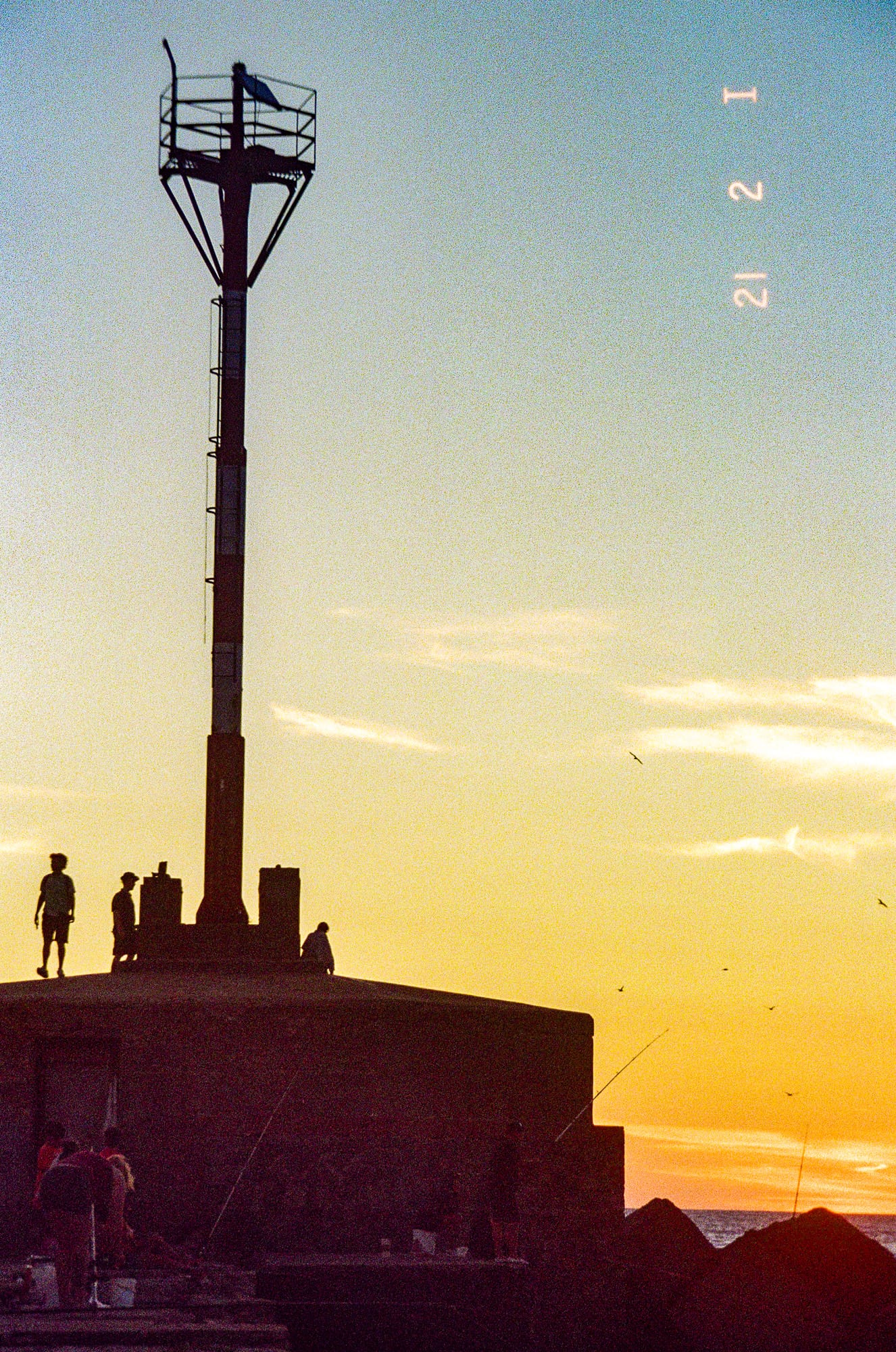 Silhouette d'une haute tour de signalisation côtière avec personnages à sa base devant un ciel orange doré au coucher du soleil, argentique couleur