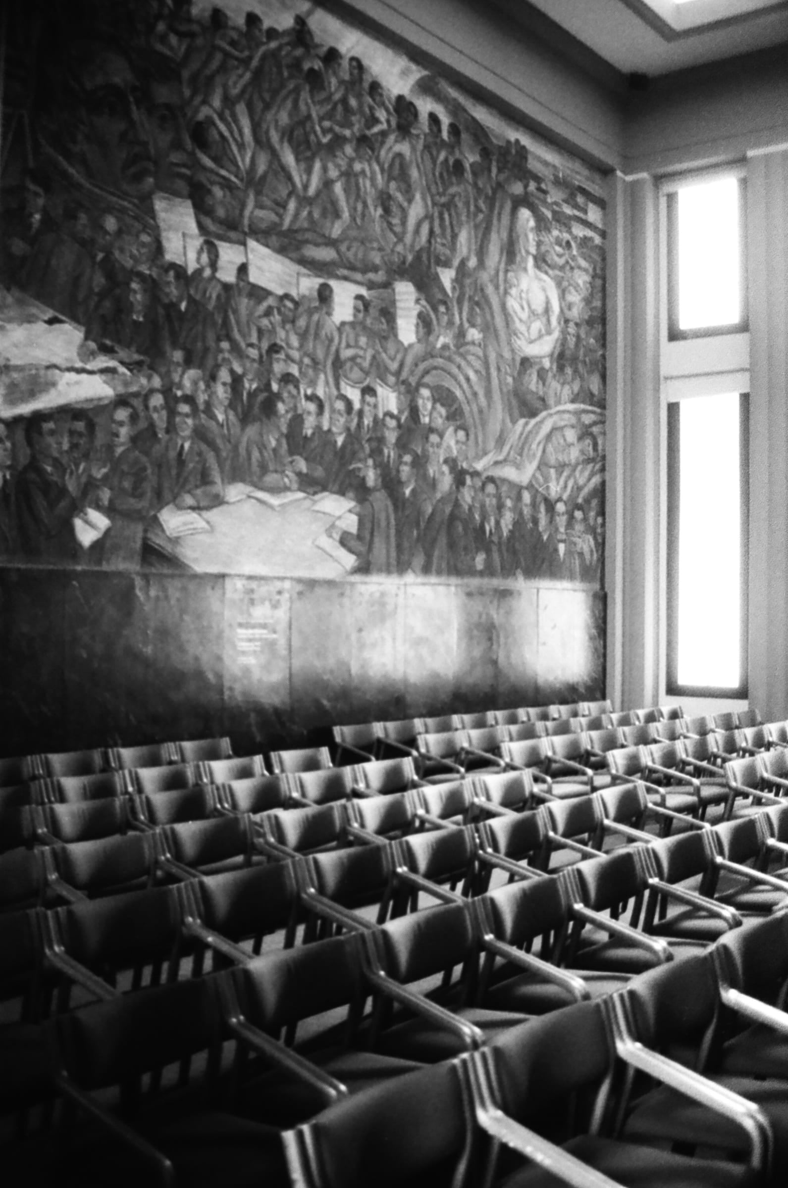 Auditorium vide aux rangées de chaises sous une grande fresque réaliste-sociale, photographie noir et blanc, Medellín