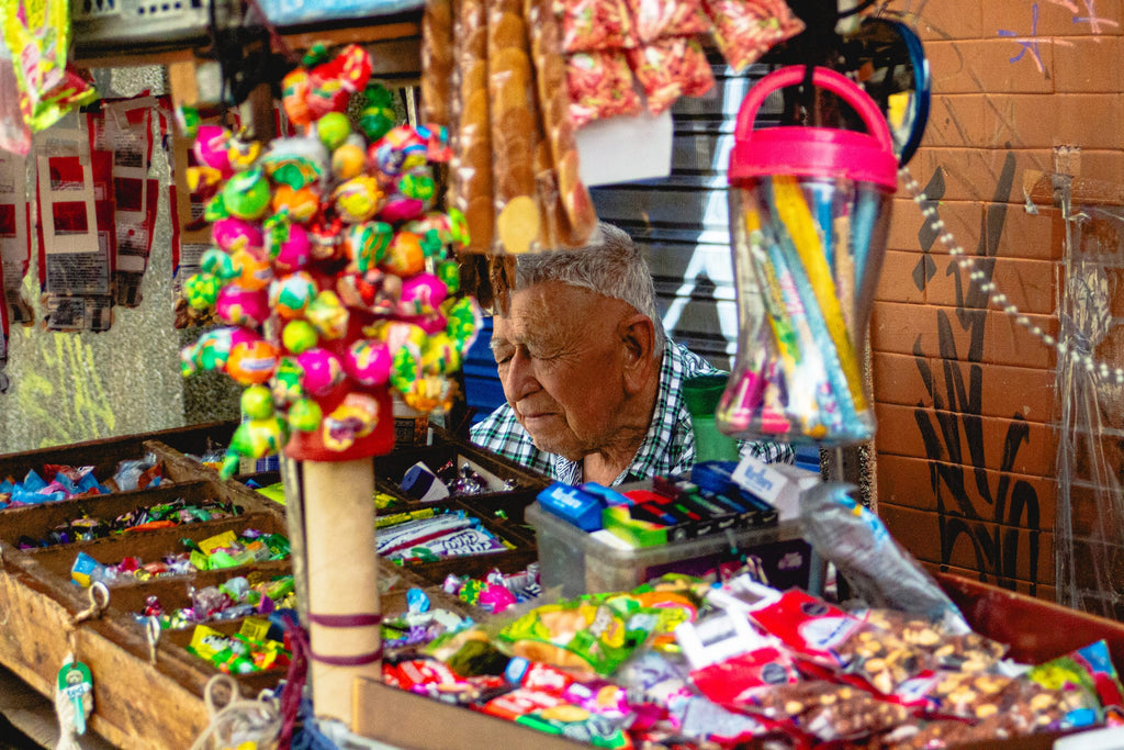 Vendeur de bonbons âgé se reposant à un étal de marché coloré débordant de sucreries et sucettes à Medellín, portrait documentaire