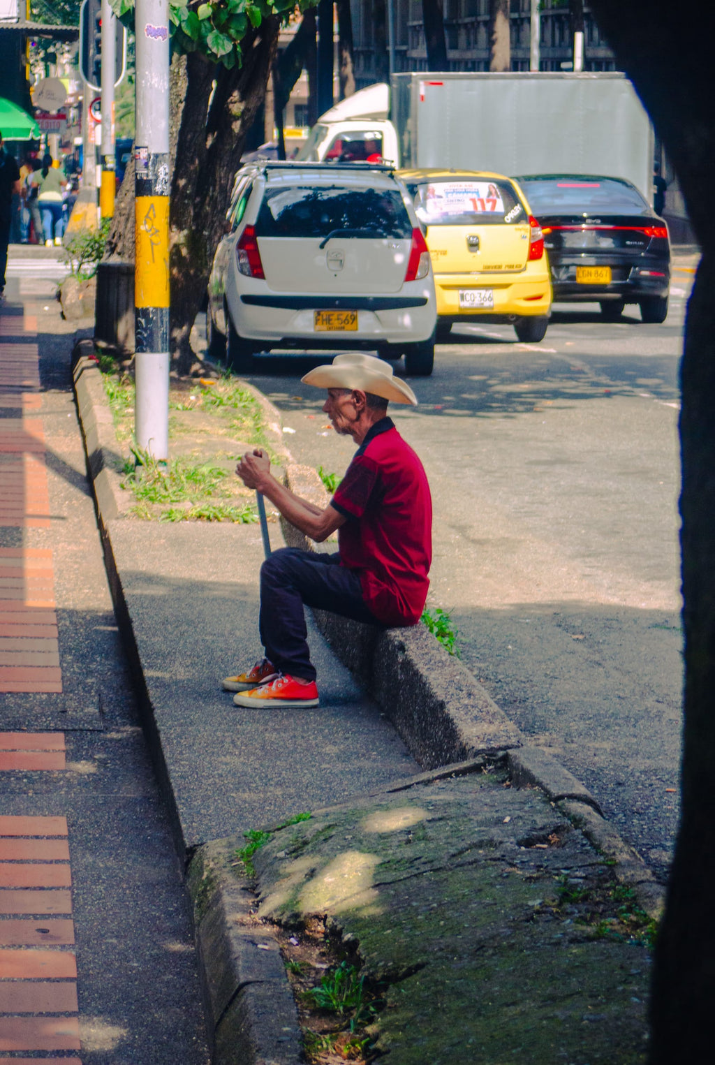 Homme âgé en chapeau de paille et chemise rouge assis sur un trottoir à Medellín, portrait de rue, documentaire couleur