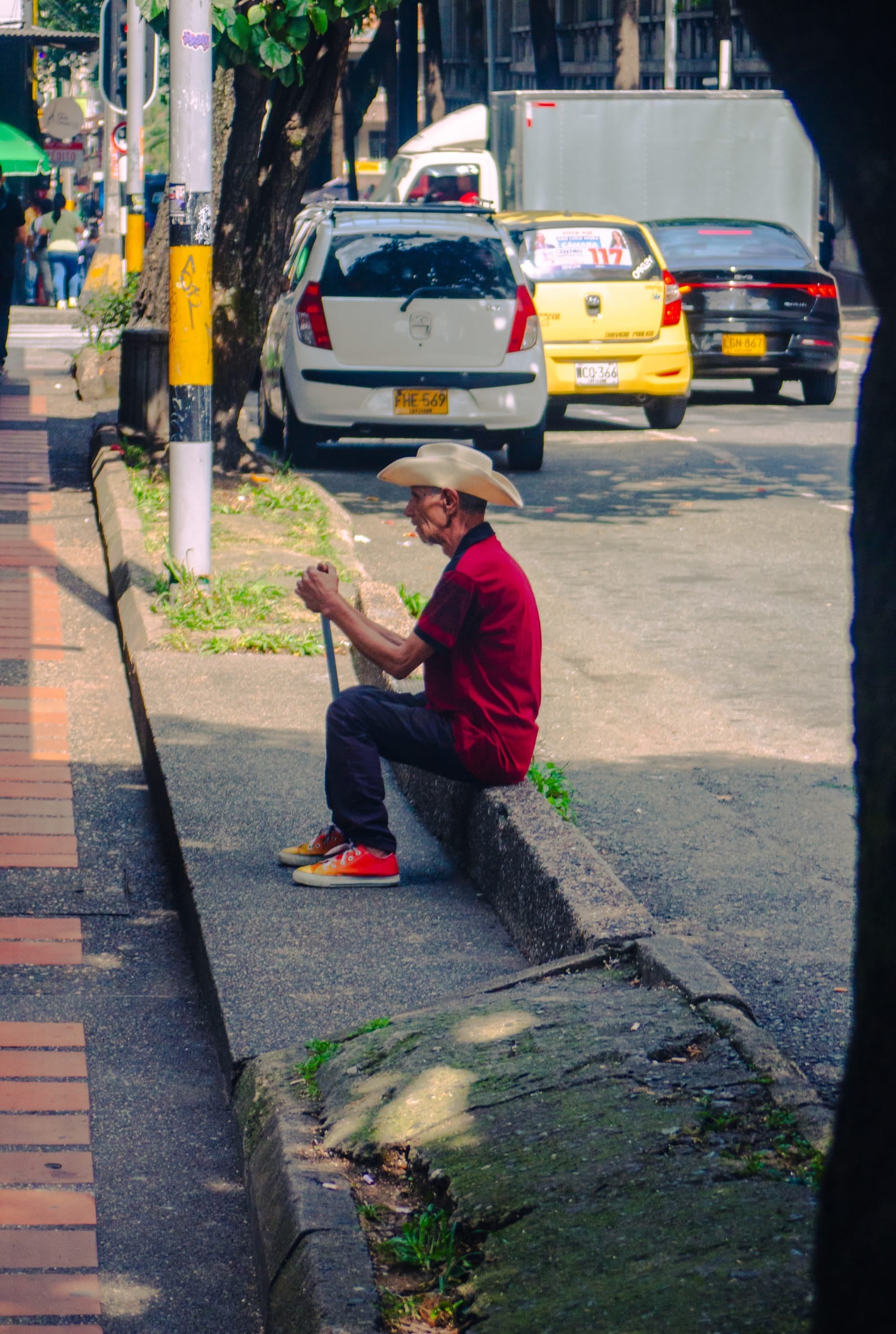 Homme âgé en chapeau de paille et chemise rouge assis sur un trottoir à Medellín, portrait de rue, documentaire couleur