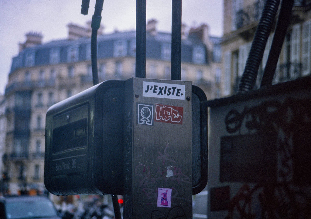 Boîtier de signalisation parisien avec sticker J'EXISTE et tags graffiti, immeubles haussmanniens flous en arrière-plan, argentique couleur