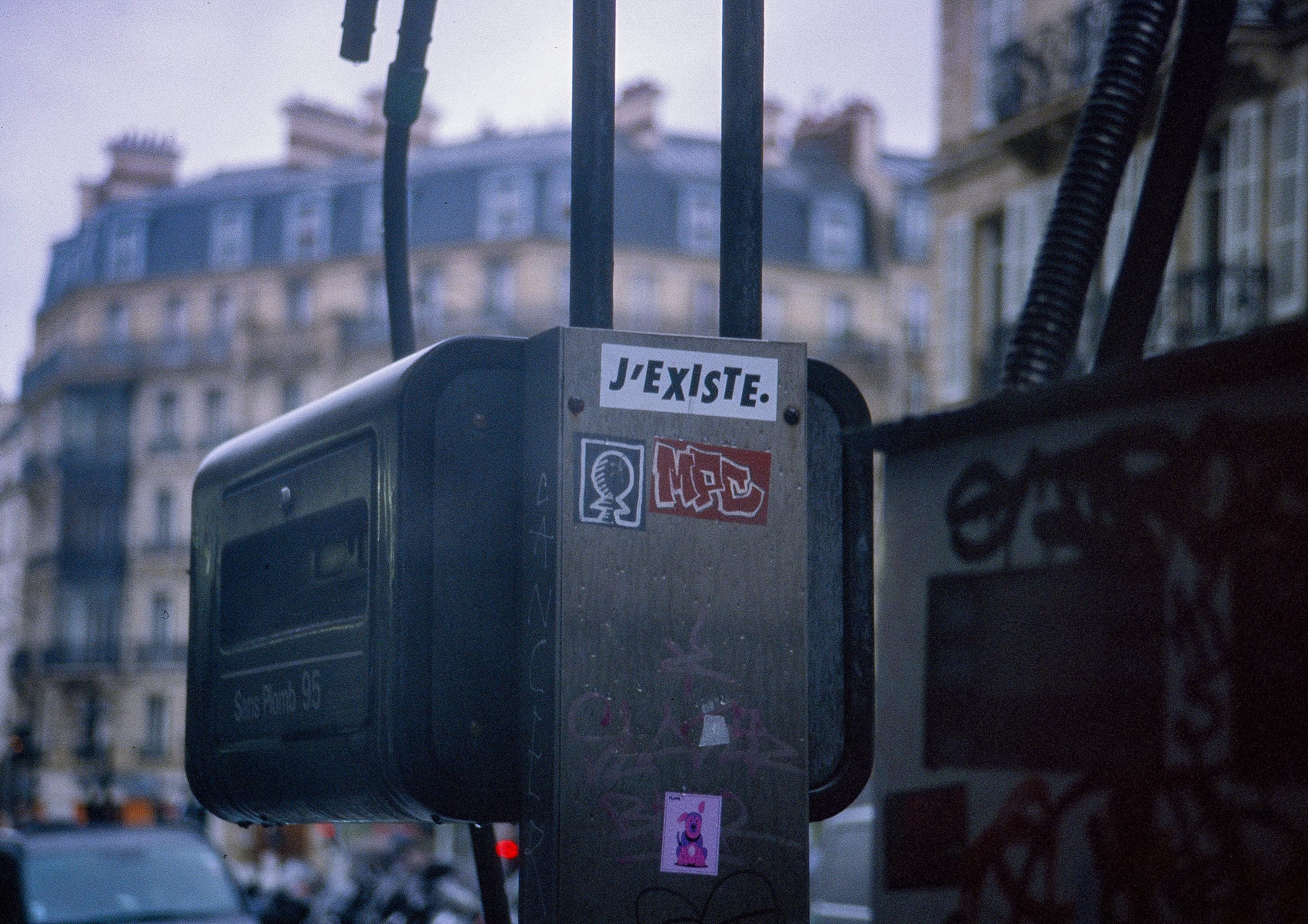 Boîtier de signalisation parisien avec sticker J'EXISTE et tags graffiti, immeubles haussmanniens flous en arrière-plan, argentique couleur