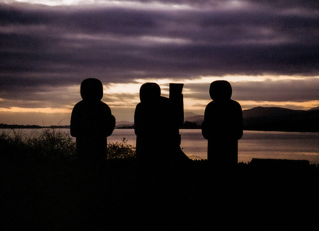 Trois figures de pierre sculptées en silhouette devant un lac patagonien violet au coucher du soleil, photographie argentique couleur