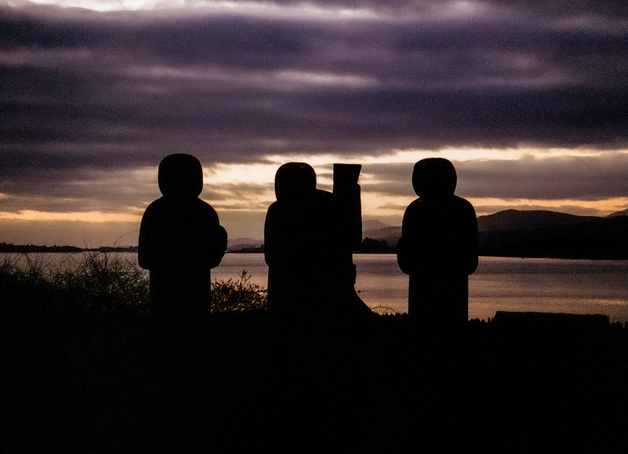 Trois figures de pierre sculptées en silhouette devant un lac patagonien violet au coucher du soleil, photographie argentique couleur